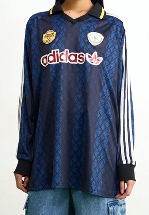 Osoba ubrana w ciemnoniebieską koszulkę Adidas z długim rękawem, z powtarzającym się wzorem liści, białe paski na rękawach oraz logo i naszywki Adidas na piersi.