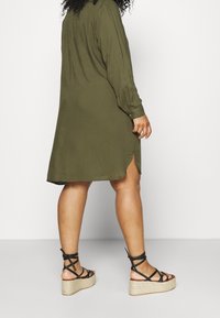 Olivgrünes, langärmliges Hemdkleid mit einem Schlitzsaum, kombiniert mit schwarzen, geflochtenen Keil-Sandalen mit Jute-Plateau.