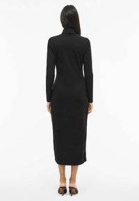 Černé šaty s dlouhými rukávy, v polodélce, s vysokým límcem, hladkou texturou a přiléhavým střihem, doplněné černými botami na vysokém podpatku.