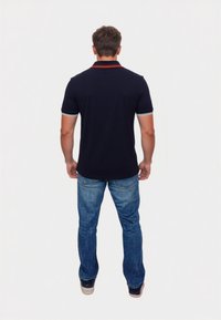 Sötétkék galléros póló piros gallérral és fehér díszítéssel, kék farmernadrággal és világos színű cipővel, hátulnézetből.