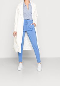 Witte lange jas over een blauw gestreepte shirt en blauwe jogger broek. Witte sneakers en een kleine witte handtas maken de outfit af.