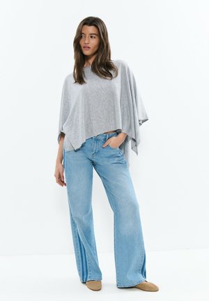 Frau steht und trägt ein lockeres graues Poncho-Oberteil, hellblaue, weit geschnittene Jeans und braune flache Schuhe vor weißem Hintergrund.