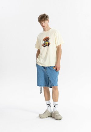 Joven con camiseta crema con un gráfico de oso de peluche, pantalones cortos de mezclilla azules, calcetines blancos con patrón y zapatillas beige con textura.