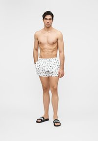 Skjorteløs mann iført hvite badebukser med mønster og svarte Moschino-sandaler, stående med hendene i lommene mot en ensfarget bakgrunn.