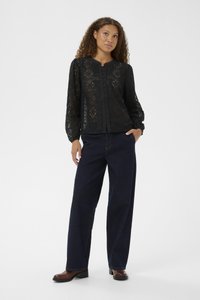 Blusa in pizzo nero con motivi testurizzati e dettagli con bottoni, abbinata a pantaloni in denim scuri a gamba larga e stivaletti marroni.