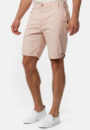 Homme portant un short rose pâle retroussé et des chaussures blanches sans lacets, debout devant un fond uni.