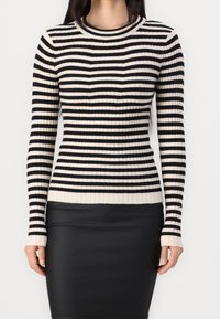 Femme portant un pull côtelé à rayures horizontales noires et blanches et une jupe noire ajustée, debout devant un fond uni.