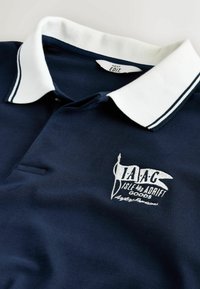 Tmavomodré polokošeľa s bielym golierom a vyšívaným logom s nápisom "I.A.A.C Isle Adrift Goods" na hrudi.