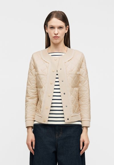 Kvinna som bär en beige vadderad jacka över en svart-vit randig skjorta och mörkblå jeans, stående mot en enfärgad vit bakgrund.