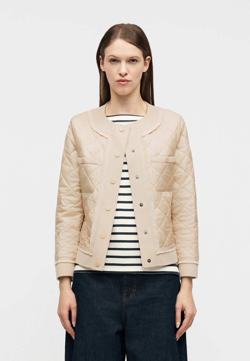 Γυναίκα που φοράει μπεζ μπουφάν με ραφές πάνω από μαύρο-άσπρο ριγέ πουκάμισο και σκούρα μπλε τζιν, στέκεται μπροστά σε λευκό απλό φόντο.