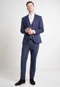 Marineblauer Anzug mit strukturiertem Design, bestehend aus einem dreiteiligen Stil, ergänzt durch ein weißes Hemd mit schwarzen Knöpfen und schwarzen Schuhen.