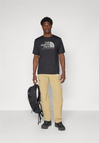 T-shirt noir à manches courtes avec le logo "The North Face", pantalon cargo beige, sac à dos noir et chaussures noires robustes.