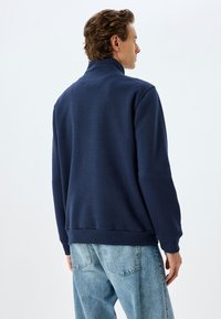 Sudadera azul marino con un cuello alto y puños acanalados. Presenta un corte holgado y una textura suave, combinada con vaqueros de un tono azul claro.