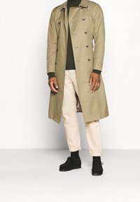 Beżowy płaszcz trench z podwójnym zapięciem i ciemnymi guzikami, nałożony na czarny golf. Jasne spodnie i czarne buty sznurowane dopełniają całość.