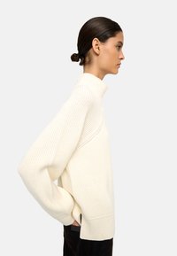 Jasna kremowa sweter z dzianiny, z wysokim kołnierzem, szerokimi rękawami i bocznymi rozcięciami, o prążkowanej fakturze i luźnym kroju.