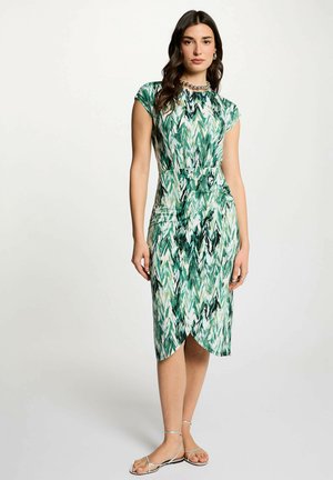 Donna in piedi che indossa un vestito verde e bianco a fantasia lungo fino al ginocchio, con maniche corte e sandali piatti con cinturini, su uno sfondo semplice.