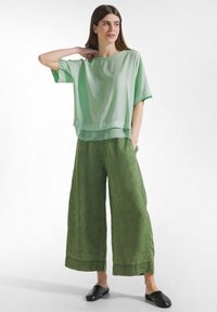 Kvinde stående med hånden bag nakken iført løs lysegrøn bluse, vidde-ben mørkegrønne bukser og sorte slip-on sko mod hvid baggrund.