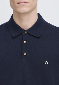 Polo din uniformă de culoare bleumarin, fabricat dintr-un material tricotat, cu guler, având trei butoane de nuanță aurie și un mic logo alb pe pieptul stâng.