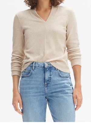 Femme portant un pull beige côtelé avec col en V rentré dans un jean bleu clair taille haute, debout, les bras détendus le long du corps.