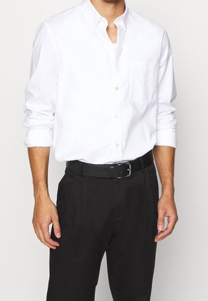 Mężczyzna ubrany w białą koszulę z długim rękawem zapinaną na guziki, wpuszczoną w czarne spodnie z czarnym paskiem, stoi na jednolitym tle.