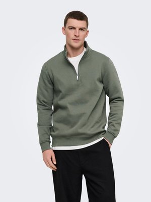 Mlad moški, ki nosi olivno zelen pulover s četrtinskim zadrgom čez belo srajco in črne hlače, stoji ob preprostem svetlem ozadju.