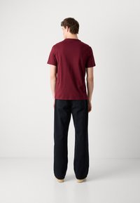 Maron T-shirt med korte ærmer, rund hals, ensfarvet design. Båret med mørke brede bukser og lyse sko, set fra ryggen.
