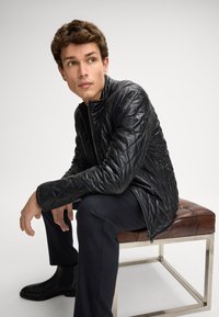 Giovane uomo seduto su una panca di pelle marrone, indossa una giacca di pelle trapuntata nera e pantaloni neri, guardando a sinistra.