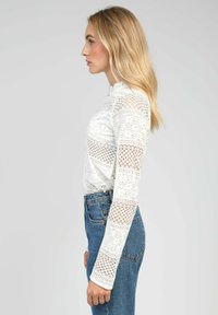 Haut en dentelle blanche à manches longues, ornée de motifs complexes et d'un col haut, associé à un jean bleu taille haute.