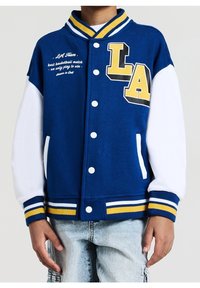 Niebiesko-biała kurtka varsity z żółtymi akcentami, z logo "LA" i napisem. Ściągacze na mankietach i u dołu, zapięcie na zatrzaski, miękki materiał.