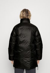 Doudoune noire avec un col montant, coupe ample et manches longues. Présente une texture lisse et brillante ainsi qu un cordon de serrage ajustable à l'ourlet.