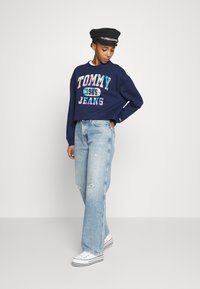 Námornícky skrátený sveter s viacfarebným logom "TOMMY JEANS 1985", skombinovaný s svetlomodrými roztrhnutými širokými džínami a bielymi teniskami.