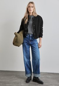Giubbotto bomber nero con bottoni argentati, maglione grigio, jeans larghi blu e stivaletti neri, con una giacca a vento verde oliva in mano.