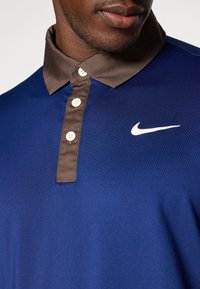 Camisa polo de Nike con un cuerpo azul marino y un cuello marrón en contraste, con tapeta de botones y logo blanco en el pecho. Textura suave.