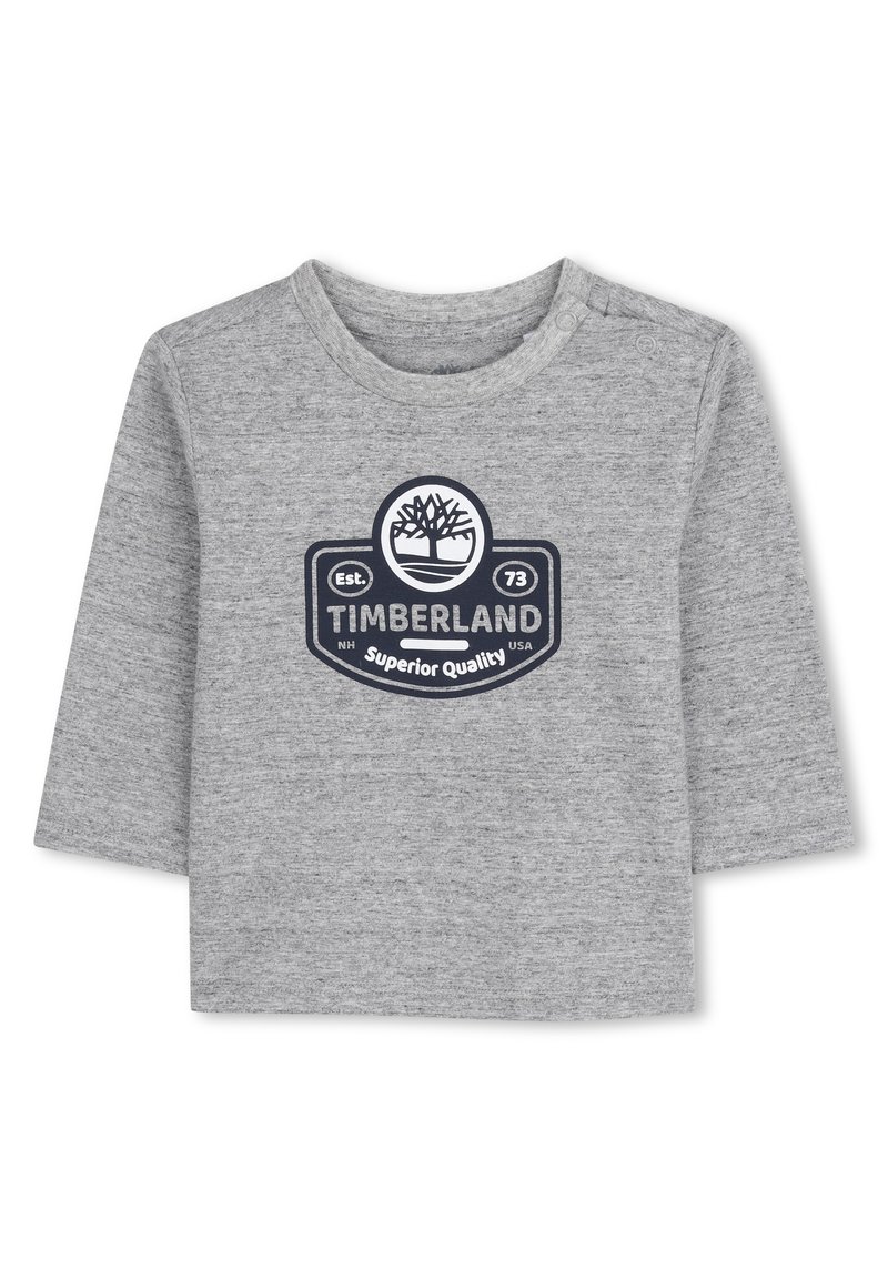 Pilkas ilgomis rankovėmis marškinas, pagamintas iš švelnios medžiagos, su apskritu Timberland logotipu tamsiai mėlynos spalvos, po kuriuo užrašai "Superior Quality" ir "Est. 73".