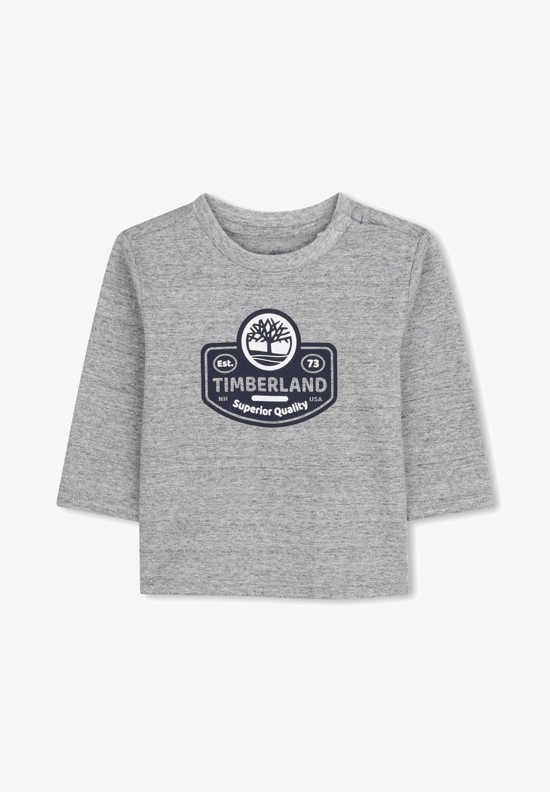 Pilkas ilgomis rankovėmis marškinas, pagamintas iš švelnios medžiagos, su apskritu Timberland logotipu tamsiai mėlynos spalvos, po kuriuo užrašai "Superior Quality" ir "Est. 73".