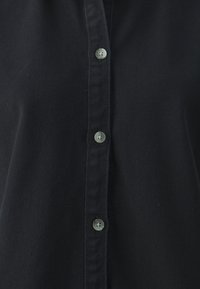 Camisa negra de botones confeccionada en tejido woven. Presenta botones grises a lo largo del placket con una textura suave y un diseño de cuello clásico.