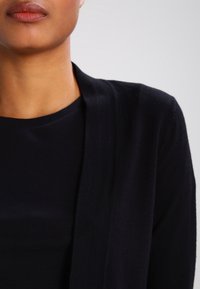 Cardigan noir avec une texture en maille douce et un design à ouverture frontale. Superposé à un chemisier noir uni, affichant une esthétique minimaliste.