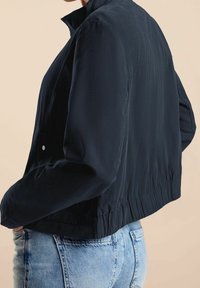 Person klädd i en svart lättviktsjacka med elastisk nederkant och ljusblå denimjeans, stående mot en beige bakgrund.