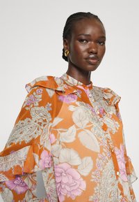 Blusa floreale arancione fluida con spalle arricciate, caratterizzata da fiori rosa e motivi intricati. Realizzata in tessuto leggero e trasparente.