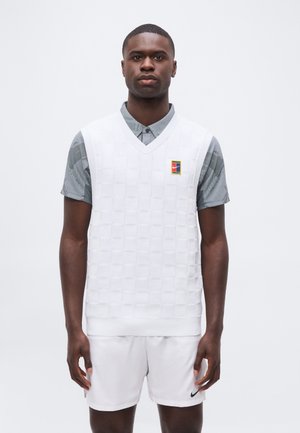 Homme portant un pull sans manches blanc à carreaux sur un polo gris à motifs et un short blanc avec des logos Nike, debout devant un fond uni.