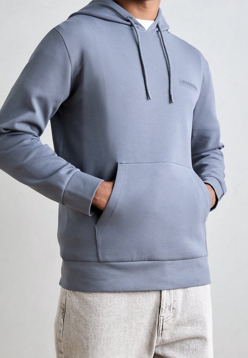 Homme portant un sweat à capuche bleu clair avec poches avant et cordons, associé à un jean de couleur claire, mains dans les poches du sweat.
