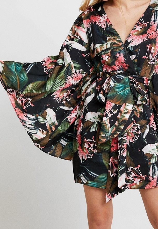 Femme portant une robe portefeuille à fleurs avec de larges manches fluides et une ceinture nouée à la taille dans des tons verts, roses, blancs et marron.