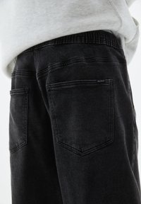 Zbliżenie osoby noszącej czarne dżinsy z elastycznym pasem i tylnymi kieszeniami, połączone z jasnoszarym swetrem.