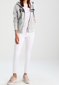 Felpa grigia con zip e testo scuro, t-shirt bianca, pantaloni slim-fit bianchi e sneakers rosa chiaro. Look casual con una trama morbida.