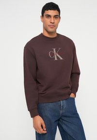 Hnědá bavlněná mikina s velkým šedým logem "CK" a červeným textem "Calvin Klein". Žebrovaný límec a manžety, volný střih.