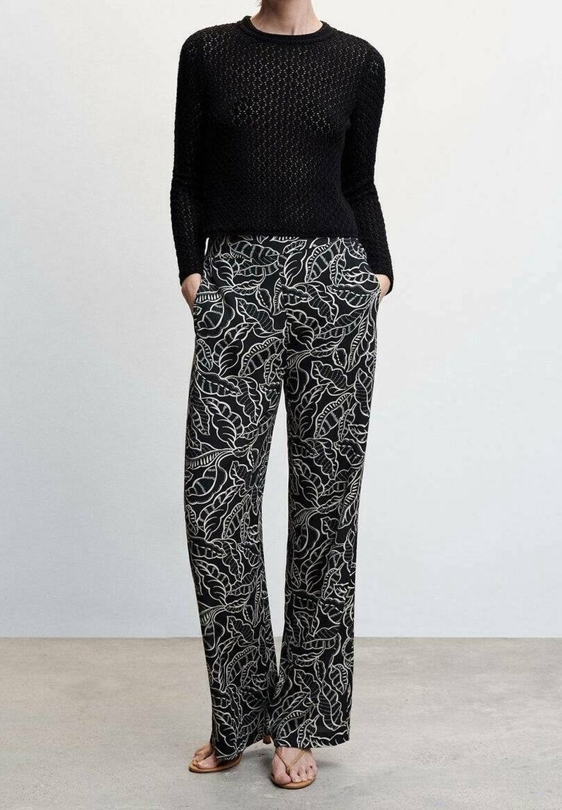 Femme portant un haut noir en maille à manches longues et un pantalon noir à jambes larges avec imprimé feuilles blanches, mains dans les poches, debout sur un sol gris.