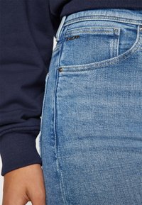 Lys blå denimnederdel med en tætsiddende form, synlig syning og et lille sort logo. Teksturen er glat med et let falme mønster.