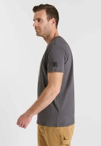 Camiseta gris de manga corta hecha de algodón suave, con un parche negro texturizado en la manga izquierda y un diseño de cuello redondo.