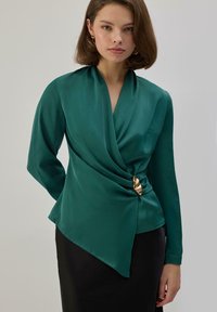 Mujer vistiendo una blusa cruzada de satén verde esmeralda con broche dorado y falda negra, de pie frente a un fondo liso gris claro.