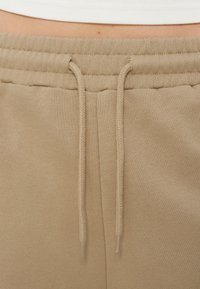 Pantalon de jogging beige avec une ceinture élastique et des cordons à l'avant, porté avec un haut blanc côtelé, laissant apparaître une petite zone de peau au-dessus de la taille.